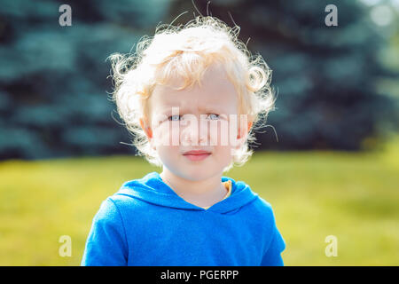 Closeup Ritratto di giovane carino Caucasian little boy ragazza con i capelli biondi e gli occhi blu con faccia buffa espressione, in felpa con cappuccio blu al di fuori nel parco a s Foto Stock