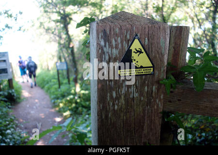 Walkers passando un cartello di avviso su un post sul sud-ovest sentiero costiero, Devon, Inghilterra, Regno Unito. Foto Stock