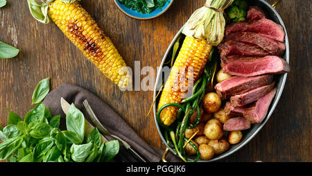 Fette di carne di manzo servita in padella con la frittura di mais, patate e fagioli verdi sul tavolo di legno con salsa di basilico, vista dall'alto Foto Stock