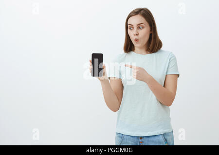 Wow Guardate cosa ho ottenuto. Impressionato entusiasta bella ragazza con breve marrone capelli labbra pieghevole in stupore rivolto verso lo schermo del dispositivo durante la detenzione e guardando lo smartphone fresco su muro bianco Foto Stock