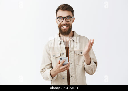 Guy entusiasti delle nuove funzioni dello smartphone che descrive gadget per gli amici dispositivo di trattenimento in mani e gesticolando con palm sorridendo entusiasta e divertito spiegando avvantaggiati del cellulare su sfondo grigio Foto Stock