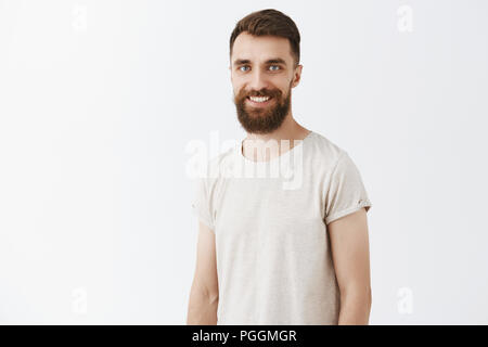 Felice e creativa dei giovani barbuto collega maschio con elegante acconciatura in bianco t-shirt casual con gioia la sorridente in piedi su sfondo grigio parlando di nuovi concetti di società la creazione di nuove idee Foto Stock