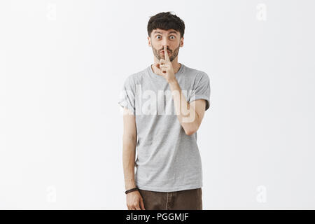 La shh faccio sorpresa. Ritratto di eccitazione emotiva di attrative giovane uomo in abito casual rendendo shush gesto con il dito indice sulla bocca come nascondere un regalo speciale o mantenendo segreti oltre il muro grigio Foto Stock