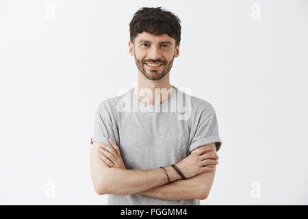 Vita-up shot di fiduciosa bella europei maturi imprenditore maschio in t-shirt tenendo le mani incrociate in auto-assicurato pongono e sorridente con assicurato sorriso piacevole su sfondo grigio Foto Stock
