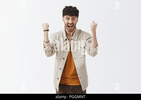 Felice e over-emotiva di calcio maschile ventola con i capelli scuri e la barba in abito elegante sollevamento rinserrata pugni, urlare con il trionfo e la felicità, tifo e sensazione di sollievo dopo aver vinto in lotteria Foto Stock