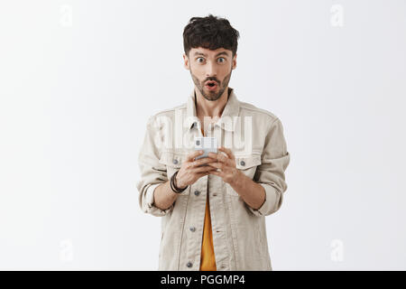 Wow check it out quale messaggio ricevuto. Ritratto di sorpreso sorpreso bello alla moda uomo con barba labbra di piegatura, tenendo lo smartphone e sguardo impressionato in telecamera raccontando cool news Foto Stock