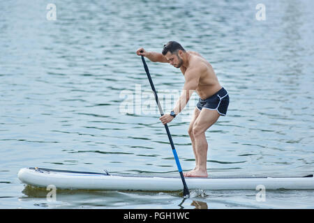 Vista laterale dell'uomo formazione con sup board, il roaming con lunghi pozza nel lago della città. Modello maschile indossare pantaloncini sport, permanente sulla grande lungo bordo, nuotare sulla superficie dell'acqua. Coltivare uno stile di vita sano. Foto Stock