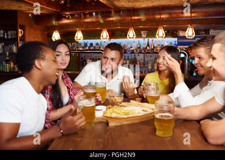 Felice gruppo di amici a bere birra alla birreria bar ristorante. Barbuto ragazzo con un bicchiere in mano, guardando all amico e sorridente, tutti intorno a godersi il tempo t Foto Stock