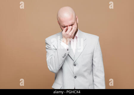 Ritratto di triste premuto di mezza età barbuto calva imprenditore della luce classico abito grigio in piedi tenendo la sua testa in basso, toccando gli occhi e pianto. in Foto Stock