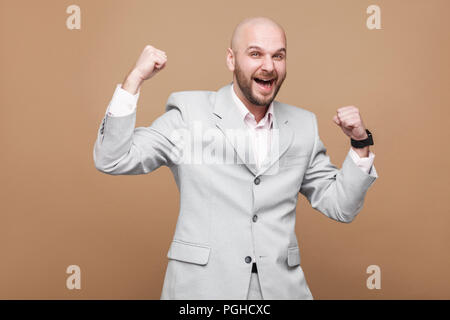 Ritratto di satisfed felice vincitore bello di mezza età barbuto calva imprenditore nel classico grigio chiaro tuta e permanente per celebrare la sua vittoria. indoo Foto Stock