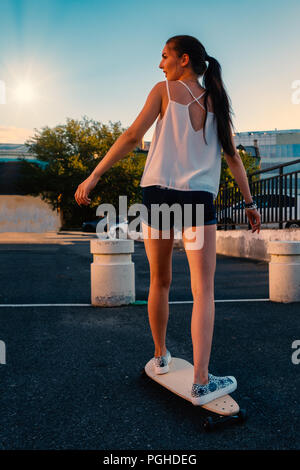 Bella ragazza skateboard di equitazione in breve gli shorts in denim, vista dal retro di fronte all inizio del cielo della sera Foto Stock