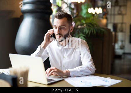 Ritratto di bello imprenditore barbuto che indossa una camicia bianca a parlare al telefono mentre si lavora a tavola in ufficio o cafe, spazio di copia Foto Stock