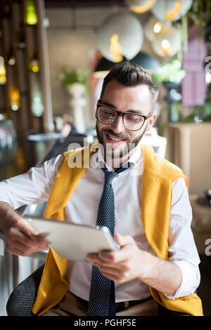Ritratto di bello barbuto imprenditore con gli occhiali seduto al banco bar con tavoletta digitale e sorridendo allegramente Foto Stock