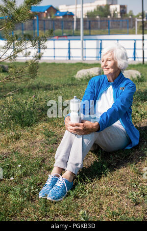 A piena lunghezza ritratto della moderna senior donna seduta di appoggio sul prato e sorridente felicemente dopo la corsa mattutina nel parco Foto Stock