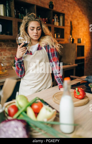 Felice casalinga in un grembiule tenendo un bicchiere di vino rosso nelle mani, cucina interno su sfondo. Cuoca prima di insalata di verdure preparazione Foto Stock