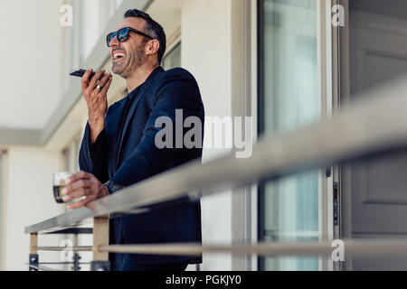 Successo imprenditore maturo in piedi nella camera di albergo balcone parlando al telefono. Allegro uomo in tuta come effettuare una chiamata dalla camera dell'albergo. Foto Stock