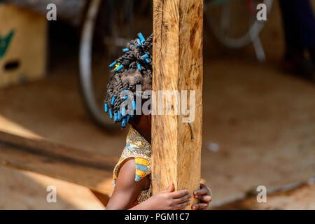 PIRA, BENIN - Jan 12, 2017: Unidentified Beninese bambina con trecce in abiti colorati si nasconde dietro il palo di legno. Il Benin i bambini soffrono di po Foto Stock