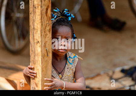 PIRA, BENIN - Jan 12, 2017: Unidentified Beninese bambina con trecce in abiti colorati si nasconde dietro il palo di legno. Il Benin i bambini soffrono di po Foto Stock