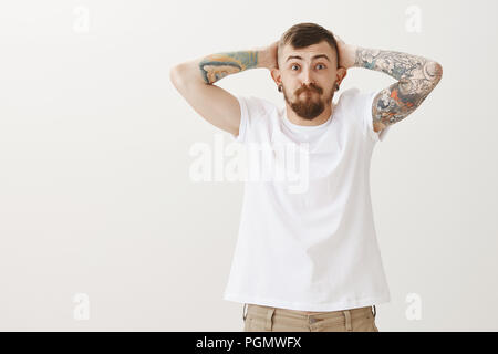 Ritratto di alimentato fino angry man cercando di tenere emozioni all'interno, busbana francese e fissando con occhi schioccato in telecamera, tenendo le mani dietro la testa, mostrando i tatuaggi sulle braccia in piedi su sfondo grigio Foto Stock