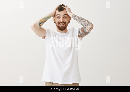 Incredibile le cose accadere. Ritratto di felice positivo giovane uomo con la barba e tatuaggi, toccando i capelli e sorridendo con gioia, sensazione eccitato e contento dopo la ricezione di notizie incredibili che cambia la vita Foto Stock