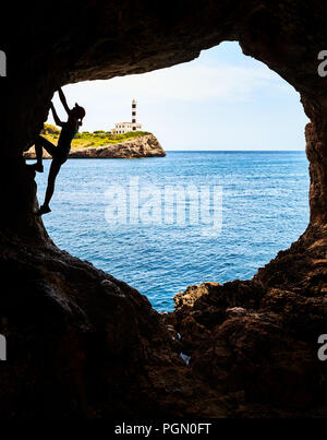 Silhouette di un atleta femminile di arrampicata in una grotta, Portocolom faro su una scogliera in background, Mallorca, Spagna. Foto Stock