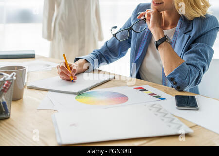 Trasformare le idee in capi di abbigliamento. Giovane e bella donna che lavorano su disegni nel suo studio vicino i vestiti appesi sulle cremagliere Foto Stock