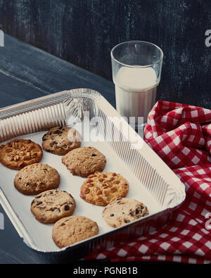 Deliziosi fiocchi d'Avena naturale biscotti al cioccolato al forno, dado, con uvetta su una teglia da forno con un bicchiere di latte e un asciugamano rosso su nero di un tavolo di legno. Foto Stock