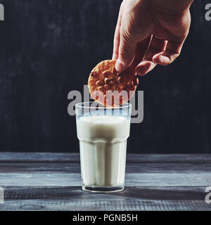 Womans dunking mano un cookie fatti in casa in un bicchiere di latte su uno sfondo nero. Foto Stock