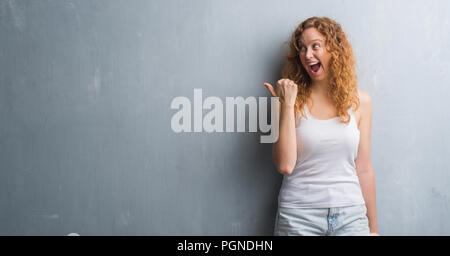 Giovane donna rosso su grigio parete grunge di puntamento e mostra con il pollice fino a che il lato con buon viso sorridente Foto Stock