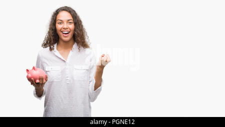 Giovane donna ispanica holding salvadanaio urlando orgoglioso e celebrando la vittoria e il successo molto eccitato, allietarla emozione Foto Stock