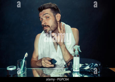 Il giovane uomo in camera da letto seduto di fronte allo specchio di graffiare la sua barba a casa. Le emozioni umane e il concetto di stile di vita Foto Stock