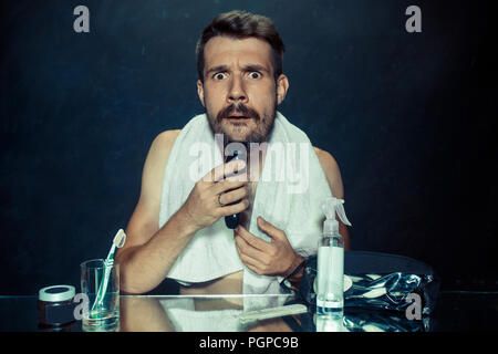 Il giovane uomo in camera da letto seduto di fronte allo specchio di graffiare la sua barba a casa. Le emozioni umane e il concetto di stile di vita Foto Stock