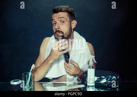 Il giovane uomo in camera da letto seduto di fronte allo specchio di graffiare la sua barba a casa. Le emozioni umane e il concetto di stile di vita Foto Stock