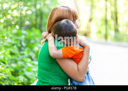 Bella donna e il suo piccolo figlio di tremore e fortemente abbracciando, sul parco della città sullo sfondo. Estate ritratto di madre e figlio per la Festa della mamma. Foto Stock