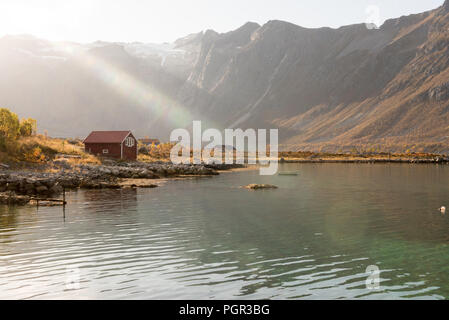 Fasci di luce con lens flare su un picco di montagna shining giù sopra una capanna sulla spiaggia in Tromso, Norvegia in una nebbiosa giornata in una scenografica landsc rurale Foto Stock