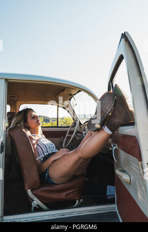 Donna addormentata gambe appoggiate sulla porta aperta dell'auto retrò e dormite sul sedile alla luce del sole Foto Stock