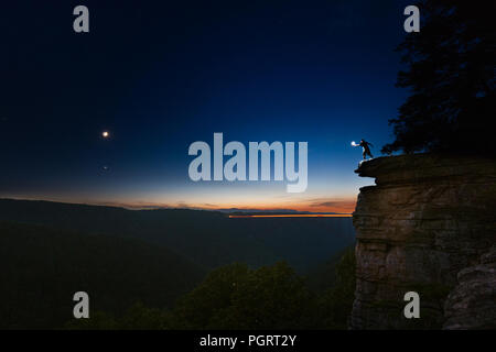 Un uomo si erge stagliano contro il cielo sul bordo di una scogliera con una mano illuminato di raggiungere attraverso un abisso di fronte alla falce di luna nella notte. Foto Stock