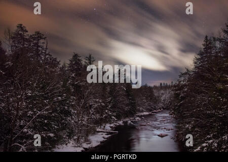 Nuvole streak attraverso il West Virginian cielo notturno passando sopra il fiume Blackwater e coperta di neve sotto i pini in questa lunga esposizione. Foto Stock