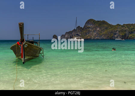 Una barca lunga vicino alla spiaggia e due snorklers off a lato con una montagna di calcare in background e cielo blu. Foto Stock