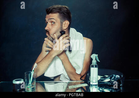 Il giovane uomo in camera da letto seduto di fronte allo specchio di graffiare la sua barba a casa. Le emozioni umane e il concetto di stile di vita Foto Stock