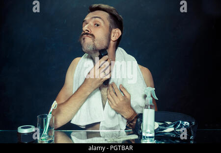 Il giovane uomo in camera da letto seduto di fronte allo specchio di graffiare la sua barba a casa. Le emozioni umane e il concetto di stile di vita Foto Stock