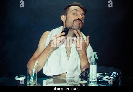 Il giovane uomo in camera da letto seduto di fronte allo specchio di graffiare la sua barba a casa. Le emozioni umane e il concetto di stile di vita Foto Stock