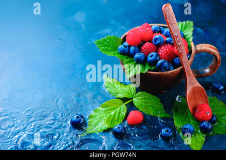 Mix assortiti di lampone e mirtillo con foglie verdi in una coppa in ceramica. Estate berry concetto raccolto su un blu scuro dello sfondo umido con gocce di pioggia Foto Stock