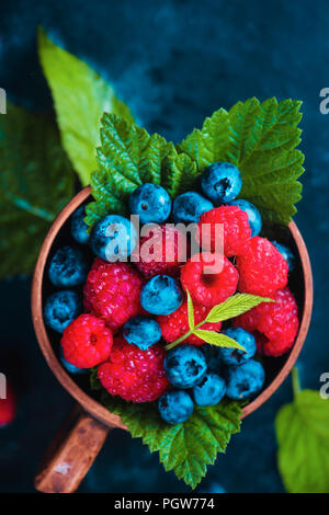 Lampone e mirtillo con foglie verdi in una coppa in ceramica. Estate berry concetto raccolto su un blu scuro dello sfondo umido con copia spazio. Vista superiore della fotografia di cibo Foto Stock