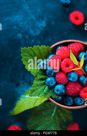 Lampone e mirtillo con foglie verdi in una coppa in ceramica. Estate berry concetto raccolto su un blu scuro dello sfondo umido con copia spazio. Vista superiore della fotografia di cibo Foto Stock