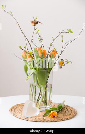 Fresh Spring mazzo di tulipani di colore arancione e verde delle foglie e due piccoli uccelli in un bel cristal vaso di vetro ed un simpatico cuore simbolo sulla scheda di paglia e t Foto Stock