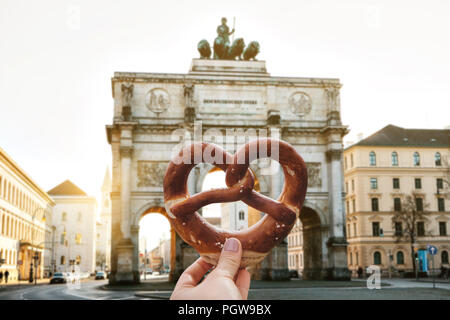 La ragazza è in possesso di una deliziosa e tradizionale tedesco pretzel nella mano contro lo sfondo della Porta Vittoria arco trionfale Siegestor a Monaco di Baviera. Germania Foto Stock