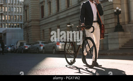 Immagine ritagliata di imprenditore a camminare su una strada di città con una bicicletta e parlando al telefono cellulare. Uomo in abbigliamento formale di andare a lavorare con ciclo utilizzando phon Foto Stock