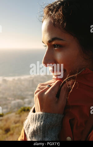 Colpo verticale del modello femminile di usura in piedi al di fuori su una scogliera e guarda lontano sorridente. Rilassata donna africana guardando una vista. Foto Stock