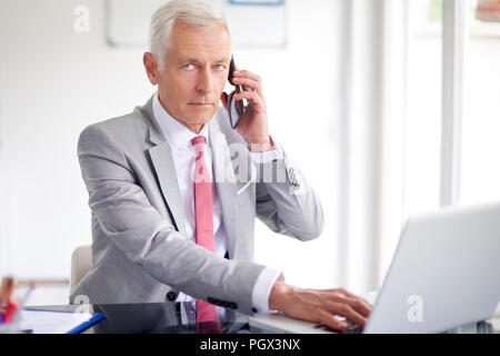 Executive senior imprenditore utilizzando il suo telefono mobile e parlando con qualcuno durante il funzionamento portatile in ufficio. Foto Stock